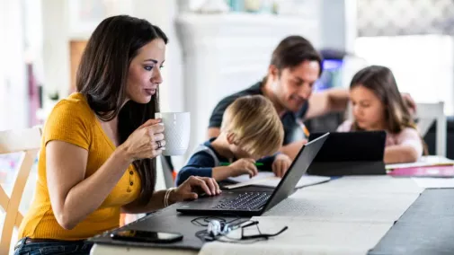 Foto einer Mitarbeiterin, die an ihrem Laptop am Familientisch arbeitet, während ihre Familie am Tisch sitzt