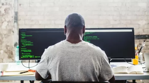 Foto der Rückansicht eines Mannes, der vor zwei Monitoren sitzt und programmiert